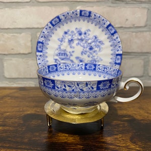 May include: A blue and white porcelain teacup and saucer set with a floral design. The cup and saucer have a gold trim. The cup is sitting on a gold metal stand.