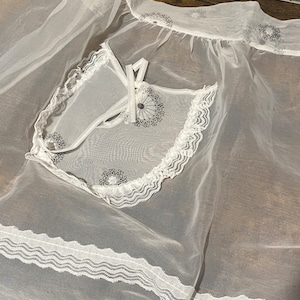 May include: White sheer apron with a lace trim and embroidered floral designs. The apron has a small pocket with a tie closure.