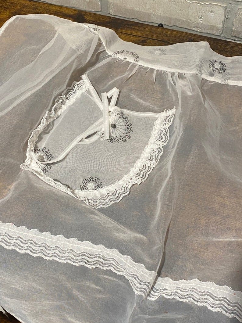 May include: White sheer apron with a lace trim and embroidered floral designs. The apron has a small pocket with a tie closure.