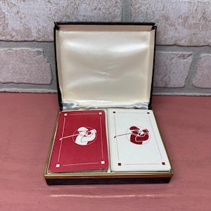 May include: A vintage playing card deck in a black box with a white interior. The cards feature a red and white design of a golfer swinging a club.