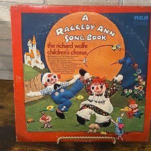 May include: A red vinyl record album cover featuring Raggedy Ann and Andy. The album is titled "A Raggedy Ann Song Book" and features the Richard Wolfe Children's Chorus. The cover art depicts Raggedy Ann and Andy dancing in a field with a castle in the background.