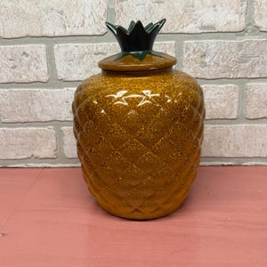 May include: A ceramic pineapple-shaped jar with a lid. The jar is a mottled brown color with a textured surface, and the lid is topped with a green pineapple crown. The jar is sitting on a pink surface.
