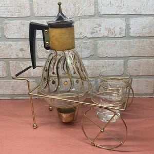 May include: Vintage glass coffee server with a gold-colored metal frame and wheels. The clear glass carafe has gold accents and a black handle. Includes matching glasses. A decorative piece for serving coffee or tea.