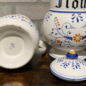 K&ouml;nnte beinhalten: Ein Set aus zwei wei&szlig;en Keramikbeh&auml;ltern mit blauen und orangefarbenen Blumendesigns. Der gr&ouml;&szlig;ere Beh&auml;lter tr&auml;gt die Aufschrift "Mehl". Der kleinere Beh&auml;lter tr&auml;gt die Nummer "4/12" auf dem Boden.