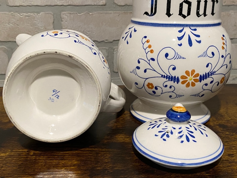 K&ouml;nnte beinhalten: Ein Set aus zwei wei&szlig;en Keramikbeh&auml;ltern mit blauen und orangefarbenen Blumendesigns. Der gr&ouml;&szlig;ere Beh&auml;lter tr&auml;gt die Aufschrift "Mehl". Der kleinere Beh&auml;lter tr&auml;gt die Nummer "4/12" auf dem Boden.
