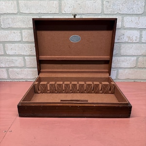 May include: Open wooden silverware chest with a brown felt interior. The chest has a hinged lid and a small metal clasp. The interior has slots for silverware and a small oval label that reads "JENEL".