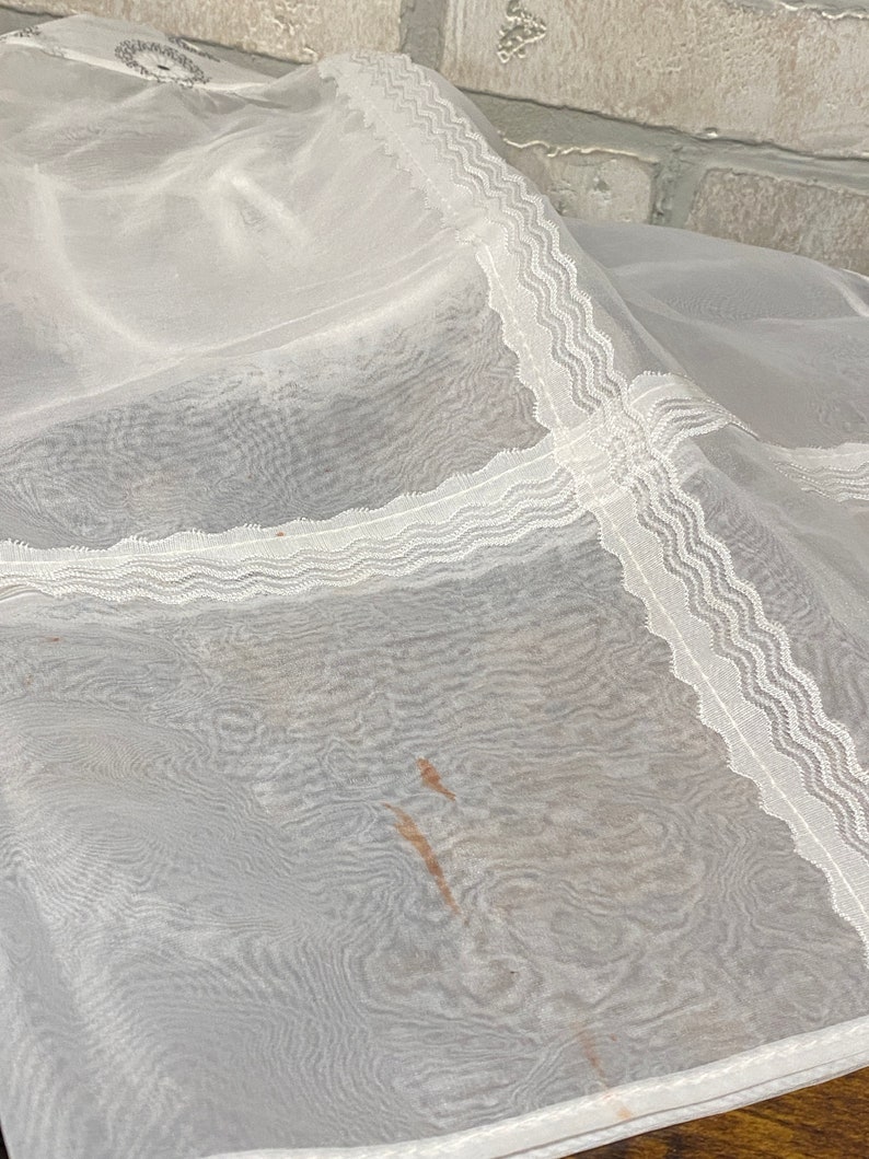 May include: White sheer fabric with a delicate lace trim. The fabric has a slight crinkle texture and a few brown stains.