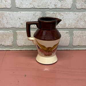 May include: A ceramic creamer pitcher featuring a two-tone design with a dark brown upper section and a cream-colored base. The front displays a graphic of a golden eagle perched above a red, white, and blue flag motif with small text below.