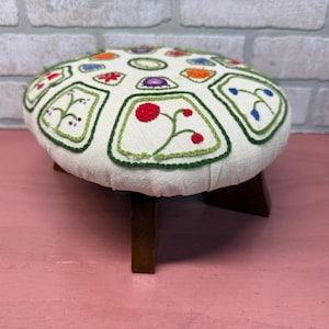 May include: A round, white footstool with a floral embroidered design. The footstool has four wooden legs.
