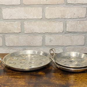 May include: Two silver metal egg poaching pans with handles. The larger pan has 12 indentations and the smaller pan has 6 indentations.