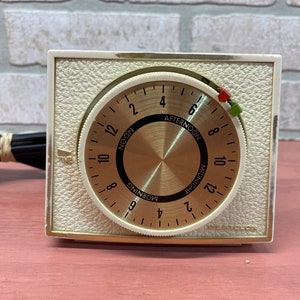 May include: A white, round, analog timer with a gold face and black markings. The timer has a dial that can be set to different times of day, including morning, noon, afternoon, and midnight. The timer is labeled "Westclox" and has a cord attached to it.