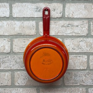 May include: Orange and red enameled cast iron pot with a black handle. The bottom of the pot has the text "De Buyer Made in Belgium".