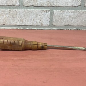 May include: A vintage wooden handled screwdriver with a metal blade. The handle is brown and has a grooved texture. The blade is silver and has a slight curve at the end.