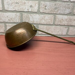 May include: A vintage copper saucepan with a long brass handle. The small pot is angled, revealing its interior. The handle extends to the right, and the pot sits on a pink surface against a brick wall.