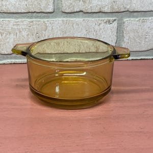 May include: Amber-colored glass casserole dish with two handles. The dish is round with a slightly flared rim. The glass has a warm, golden hue, and the dish appears to be vintage. The background is a pink surface and a brick wall.