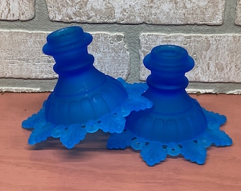 May include: Two blue glass candle holders with a lacy design around the base. The holders are frosted and have a vintage look.