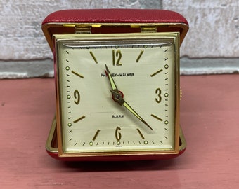 May include: A red leather-covered alarm clock with a white face and gold accents. The clock face reads "12", "3", "6", and "9" with the words "PHINEY-WALKER" and "ALARM" printed below the 12.