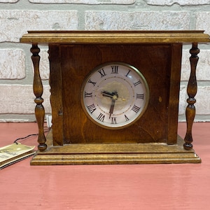 May include: A vintage wooden clock with a white face and black Roman numeral hour markers. The clock is in a wooden frame with decorative pillars on each side.