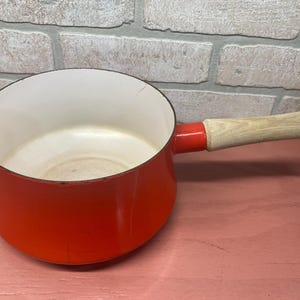 May include: A red enamel saucepan with a wooden handle. The saucepan has a white interior and is made of cast iron.