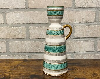May include: A white ceramic candle holder with a green and gold geometric pattern. The holder has a handle and a wide base.
