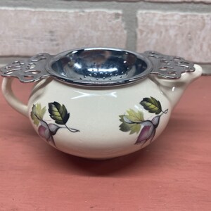 May include: A white ceramic teapot with a floral design and a silver strainer. The teapot has a handle and a spout.