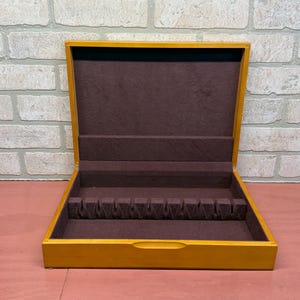 May include: A rectangular wooden storage box with a natural light brown finish and a hinged lid. The interior is lined with dark brown felt and features a molded insert with twelve individual slots designed for organizing flatware or utensils.