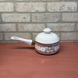 May include: White enamel saucepan with a lid and a long handle. The saucepan has a red and white star pattern and a graphic of two chefs. The handle and lid knob are white.