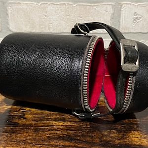 May include: A black leather cylindrical case with a zipper closure and a red lining. The case has a strap and a metal buckle.