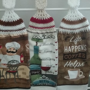 May include: Three hanging kitchen towels with crocheted tops. One features a chef, another a wine bottle with "Reds" text, and the last says "Life Happens Coffee Helps" with a coffee cup. The towels have brown, white, and red accents.
