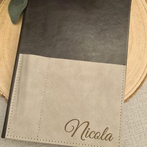 May include: A personalised notebook with a dark grey cover and a light grey pocket. The name "Nicola" is printed in a cursive font on the pocket. The notebook is on a wooden surface with pink flowers.