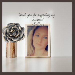 May include: A decorative arrangement featuring a grey and gold flower sculpture in a brown and beige striped vase. A framed photograph of a woman is next to the flower. The text reads, "Thank you for supporting my business! -Brittney."