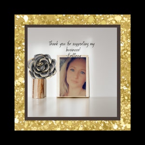 May include: Thank you for supporting my business! -Brittney. A framed photograph of a woman with blonde hair and blue eyes. A grey and gold decorative flower sits on a wooden table.