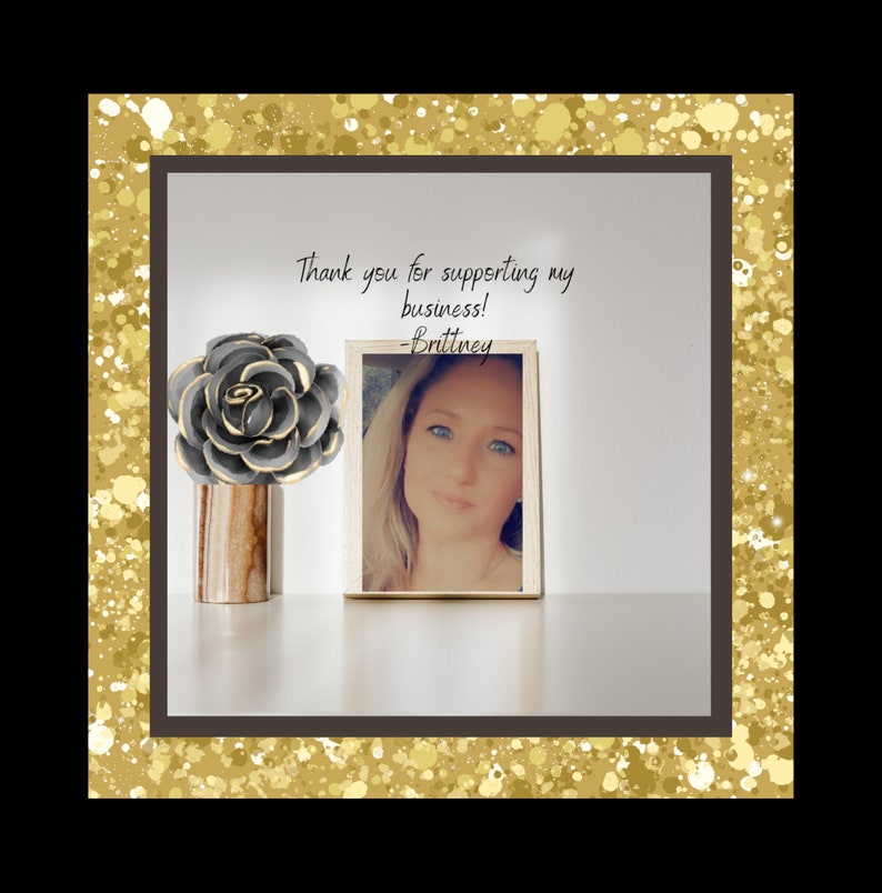 May include: Thank you for supporting my business! -Brittney. A framed photograph of a woman with blonde hair and blue eyes. A grey and gold decorative flower sits on a wooden table.