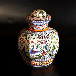 May include: A decorative ceramic jar with a lid, featuring a detailed floral and bird design. The jar has a white base with colorful patterns, including red, blue, and green accents. The lid mirrors the design, creating a cohesive look.