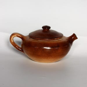 Può includere: Una teiera in ceramica marrone con un coperchio. La teiera ha un corpo arrotondato e un piccolo beccuccio. Il coperchio è leggermente bombato e ha un piccolo pomo in cima.