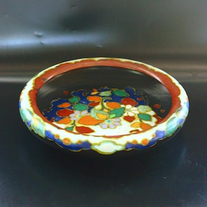 May include: A shallow, circular ceramic bowl with a black interior and exterior. The rim features a scalloped edge painted in red, blue, and white. The center of the bowl displays a vibrant floral pattern in shades of orange, green, blue, and white.