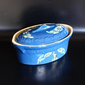 May include: An oval blue ceramic casserole dish with a lid. The dish has a cream-coloured rim and is decorated with white and yellow floral designs and a stork. The lid has a handle.