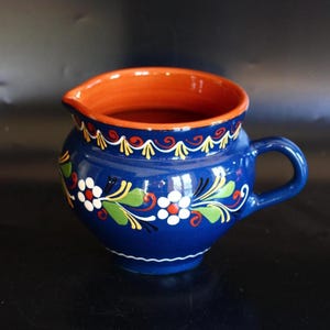 May include: A blue ceramic pitcher with a handle and a pouring spout. The pitcher has a vibrant orange interior and is decorated with white, red, and green floral patterns. The rim is adorned with a decorative yellow and white design.