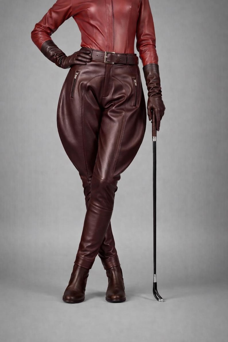 May include: A burgundy leather outfit featuring a fitted shirt, high-waisted breeches with zipper pockets, gloves, and boots. A matching belt cinches the waist. A riding crop is held in the right hand. The ensemble is set against a gray backdrop.