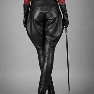 May include: Black leather riding pants with a high waist and fitted legs. The outfit includes a red leather jacket, black gloves, and a riding crop. The model is standing with legs crossed, and the background is a solid gray.