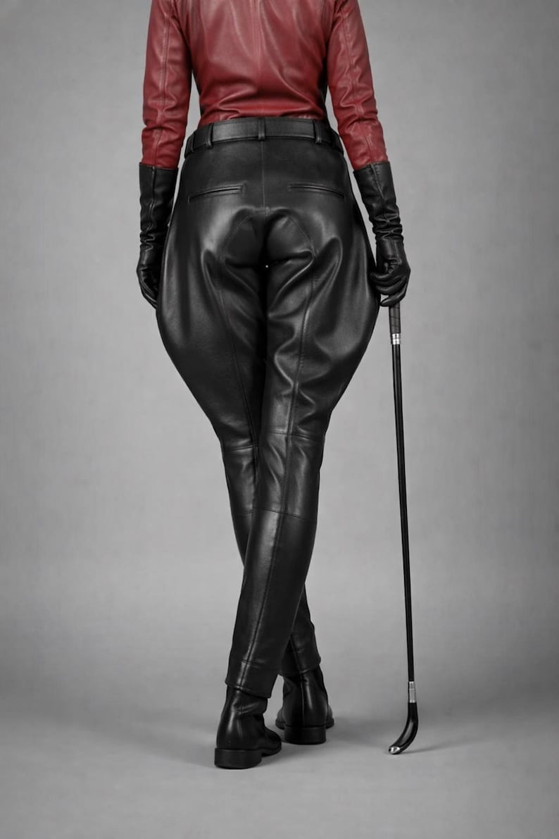 May include: Black leather riding pants with a high waist and fitted legs. The outfit includes a red leather jacket, black gloves, and a riding crop. The model is standing with legs crossed, and the background is a solid gray.
