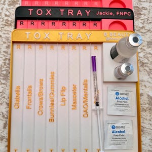 May include: A stack of three white and colored TOX TRAY organizers. The top tray is pink and white, the middle is black and pink, and the bottom is white and gold. The bottom tray contains a syringe, vials, and alcohol prep pads. Text includes "Glabella" and "DAO/Mentalis".
