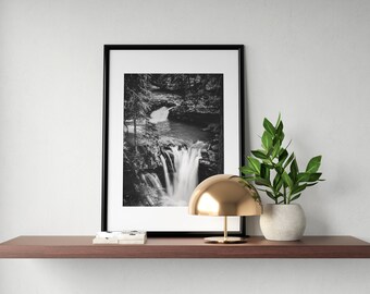 Könnte beinhalten: Ein Schwarzweißfoto eines Wasserfalls in einem Wald, gerahmt in einem schwarzen Rahmen, steht auf einem Holzregal mit einer goldenen Lampe und einer Topfpflanze.