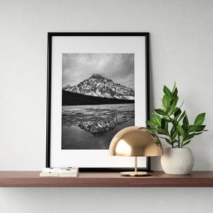 May include: Black and white framed photograph of a snow-covered mountain reflected in a lake. The photograph is displayed on a brown shelf, alongside a gold lamp and a potted plant in a white pot.