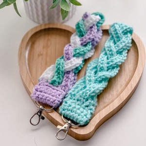 May include: Two braided crocheted keychains with silver key rings. One keychain is white, light green, and purple. The other keychain is light blue.
