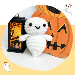 May include: A white crocheted ghost with a friendly face sits next to a glittery orange pumpkin with a black jack-o'-lantern face. The ghost has pink cheeks and black eyes. The pumpkin is decorated with glitter.