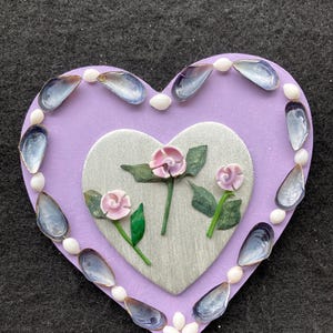 May include: A heart-shaped decorative piece with a lavender background. It features a silver heart in the center with pink roses and green leaves. The border is adorned with blue mussel shells and small white beads, creating a unique coastal design.
