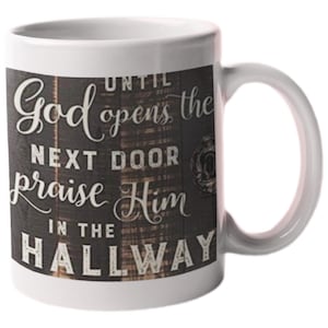 May include: White ceramic coffee mug with a black and white design. The design features a woodgrain background with the text "Until God opens the next door praise Him in the hallway".