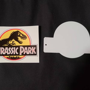 May include: A white sticker with a black dinosaur silhouette and the text "Jurassic Park" in red letters. The sticker is next to a white blank sign with two holes.