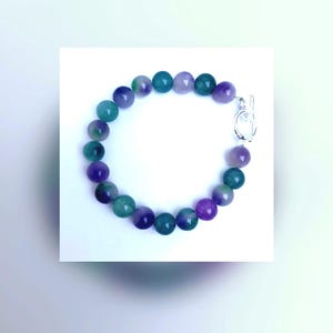 Op de afbeelding: Een kralenarmband met afwisselend ronde, gepolijste stenen in tinten dieppaars en bosgroen. De armband wordt vastgemaakt met een zilverkleurige metalen knevelsluiting, bestaande uit een ronde ring en een T-vormige staaf.